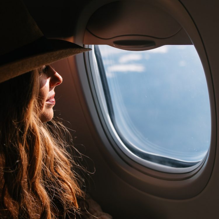 A female tourist with a hat looking out the airplane window, gazing at the blue sky and clouds.