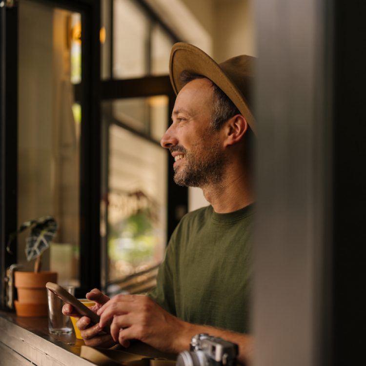Portrait of a mid adult man who enjoys warm cup of coffee in a local cafe on a beautiful autumn day, using his mobile phone while sipping coffee